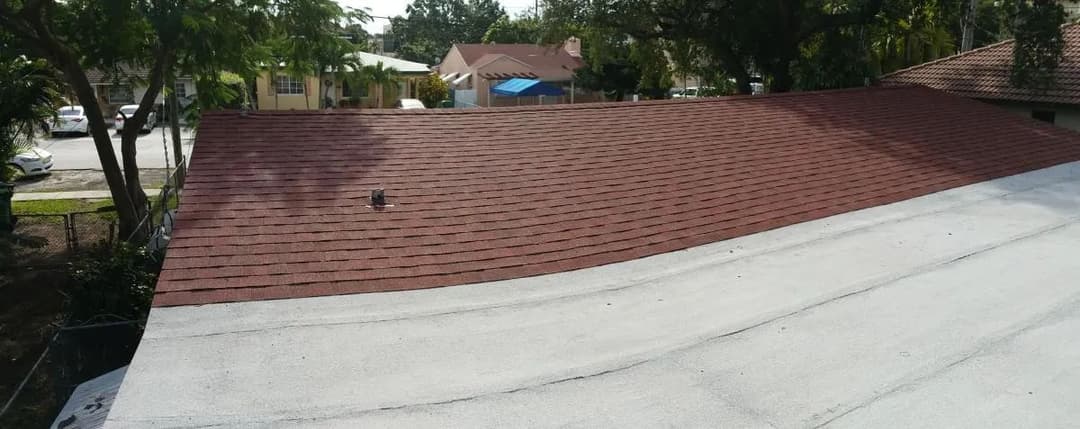 Aerial view of a residential roof with red shingles and flat white section, surrounded by greenery.