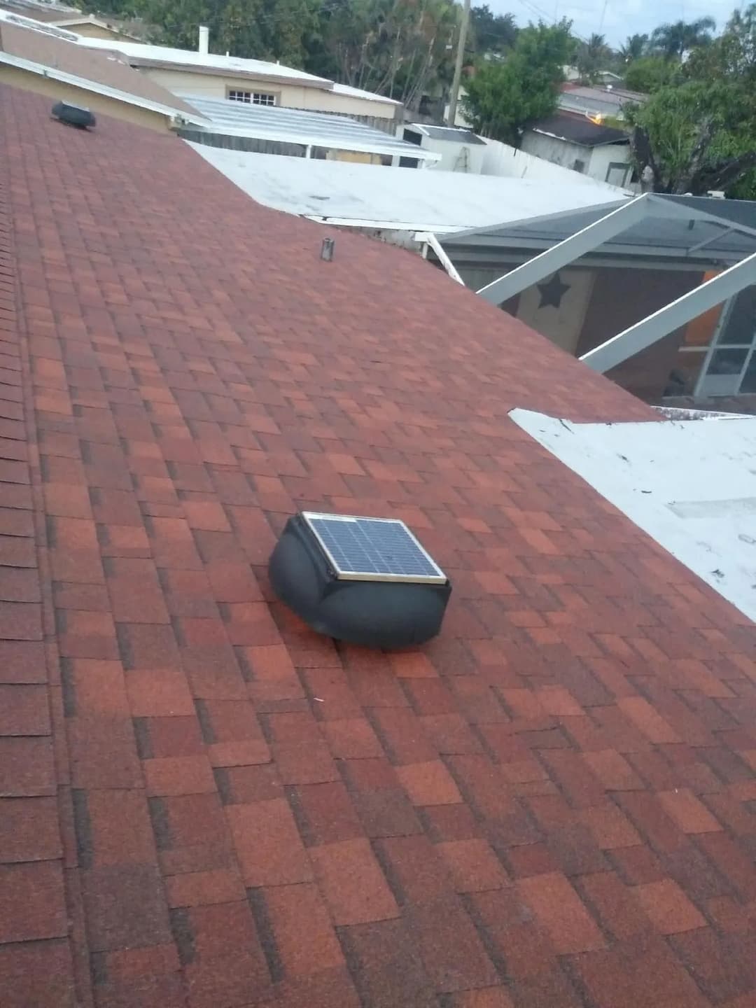 Solar attic fan on a red shingle roof with nearby flat rooftop and greenery in the background.