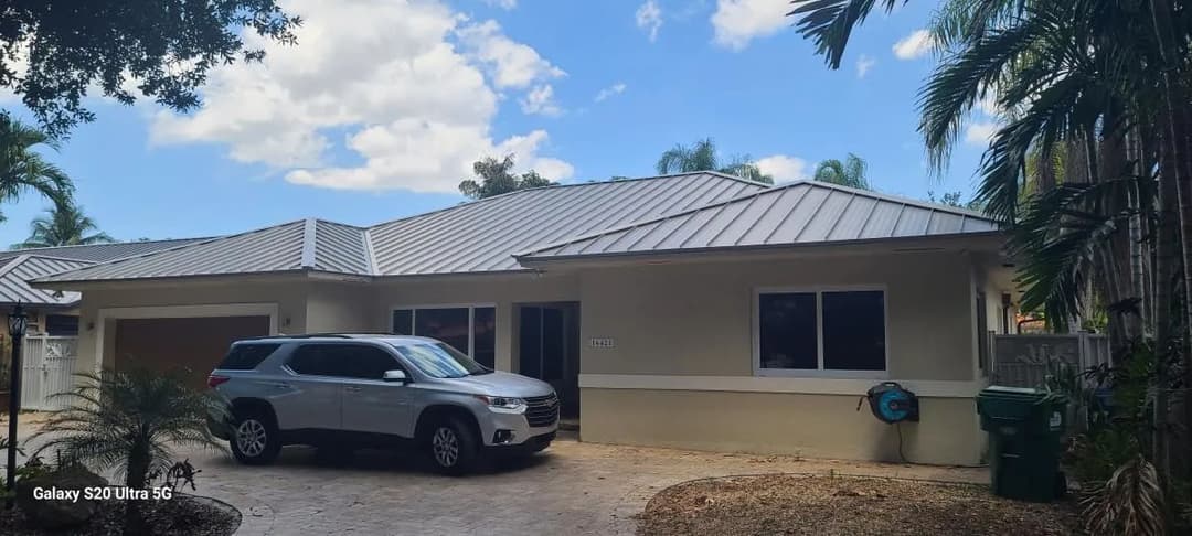 Modern single-story house with gray metal roof and parked SUV in driveway, surrounded by palm trees.