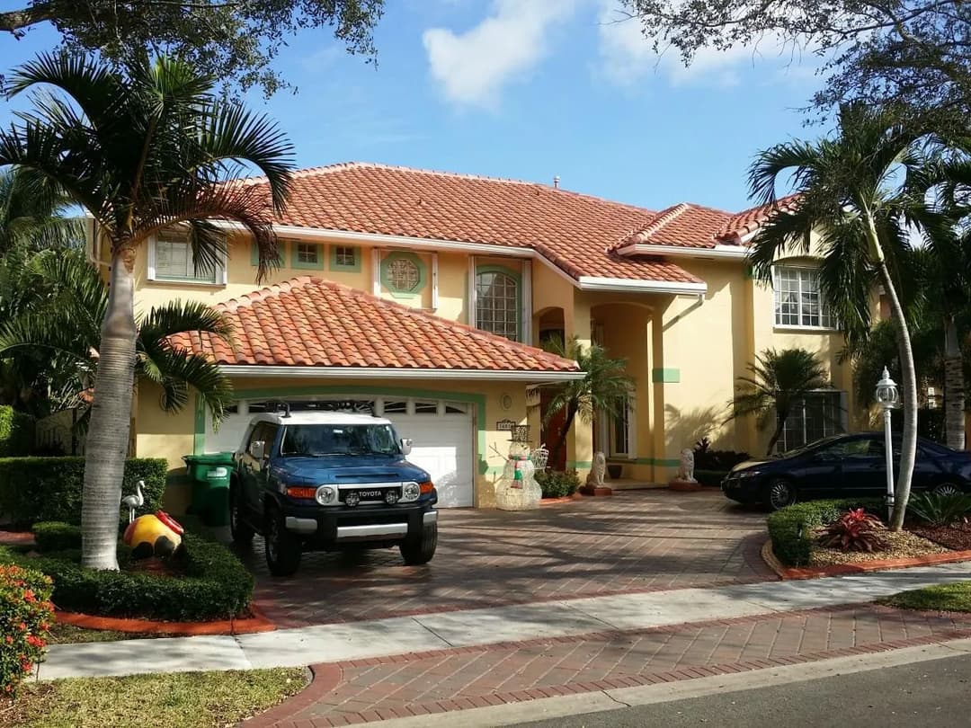 Two-story yellow house with red-tile roof, surrounded by palm trees and parked cars in driveway.