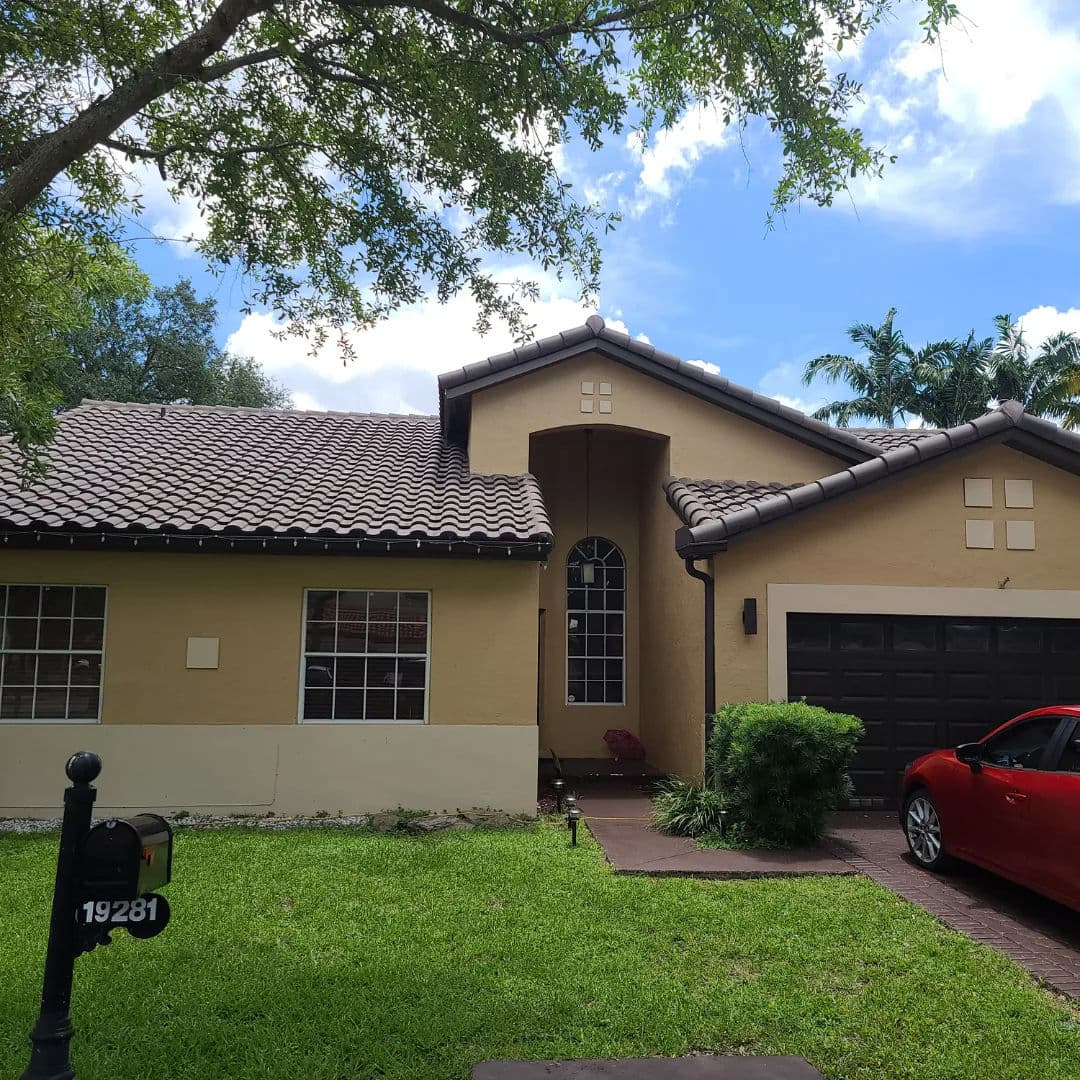 Single-story suburban home with a tile roof, red car, and lush green lawn under blue sky.